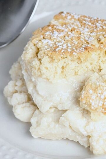 A piece of homemade vanilla pudding cake topped with powdered sugar on a white plate next to a spoon.