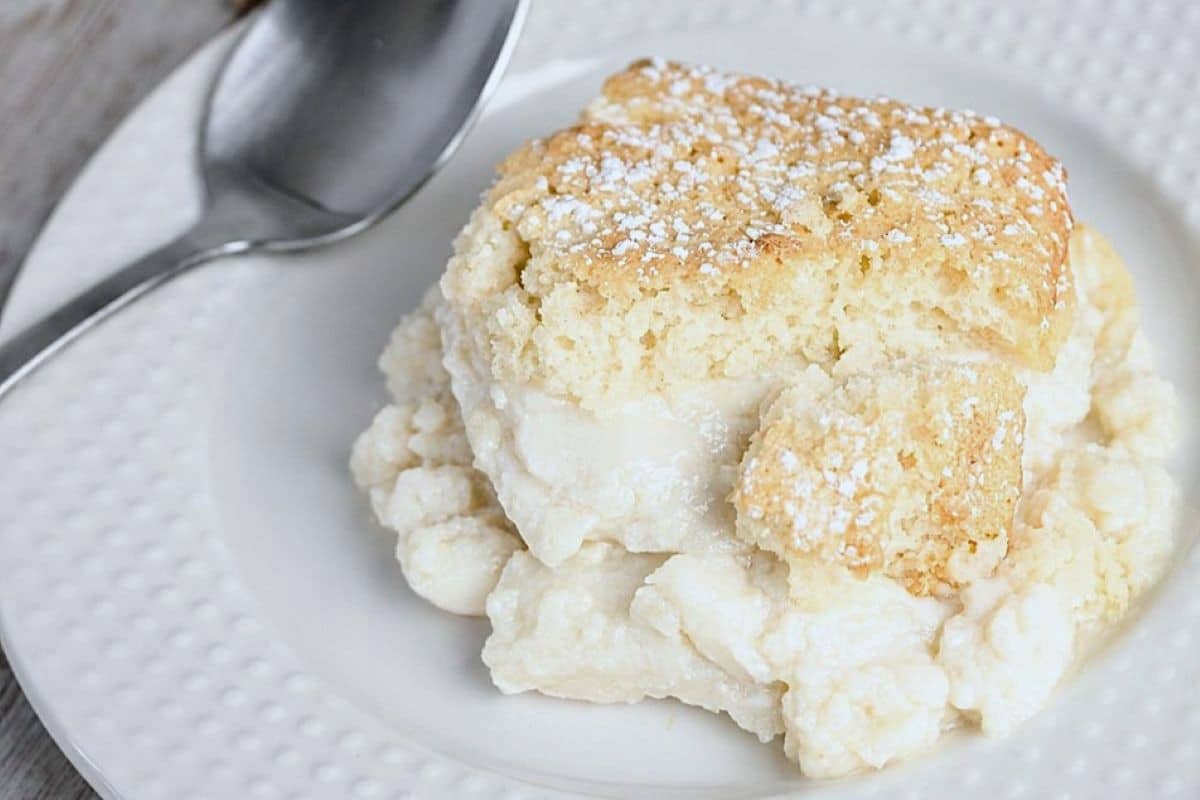 A piece of homemade vanilla pudding cake topped with powdered sugar on a white plate next to a spoon.