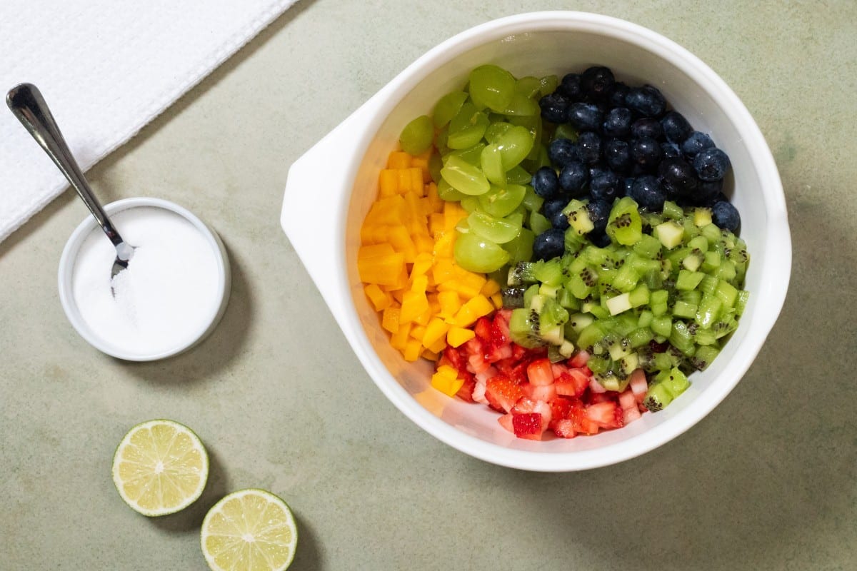 A white bowl filled with fresh fruit salsa containing diced mango, green grapes, blueberries, chopped kiwi, and diced strawberries, with a small dish of sugar, a spoon, and two lime halves nearby.