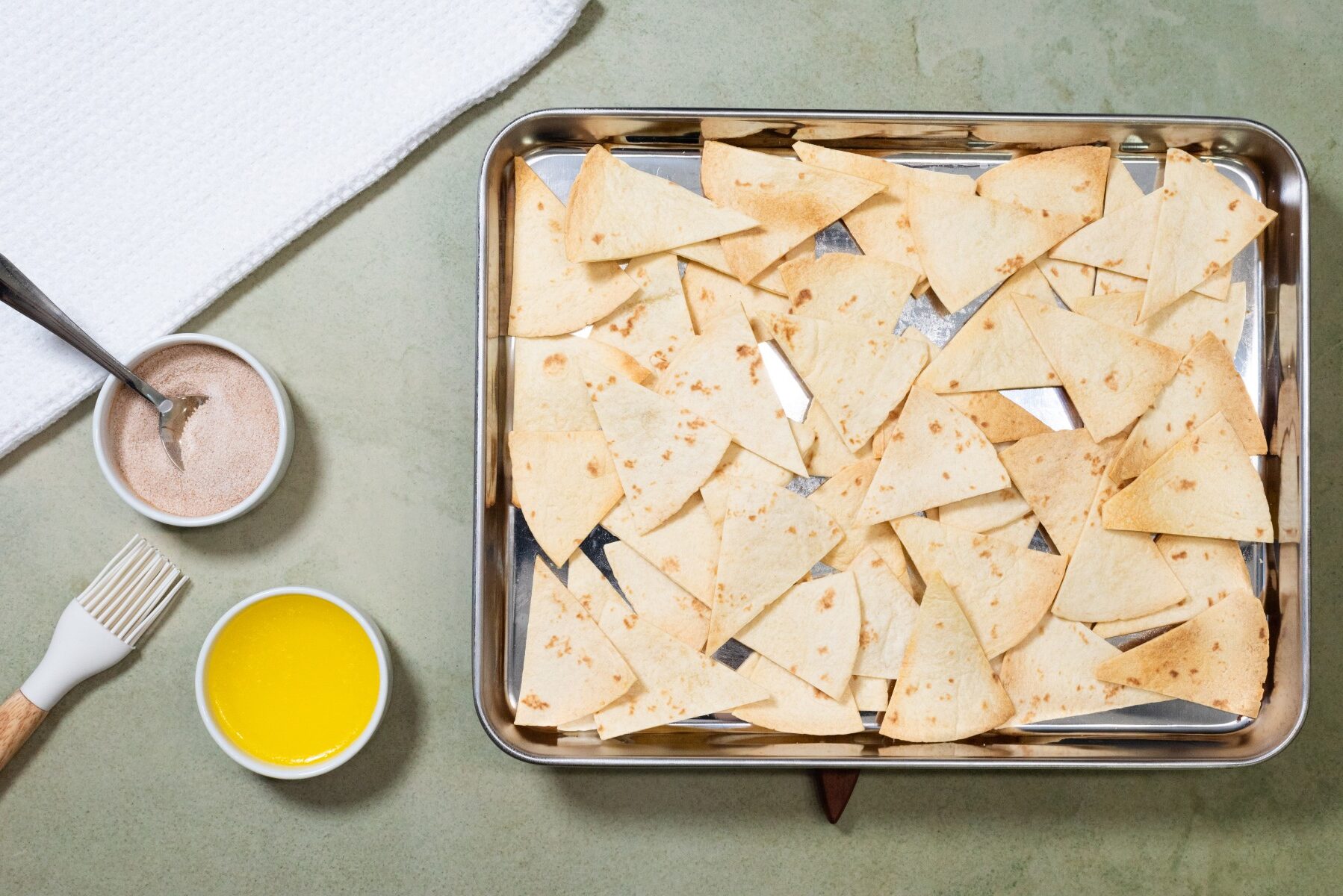 Baking sheet filled with triangular tortilla chips, with small bowls of cinnamon sugar and melted butter nearby for fresh fruit salsa.