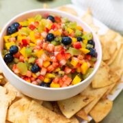 A bowl of fresh fruit salsa made with diced strawberries, mango, kiwi, and blueberries, served with a side of cinnamon sugar tortilla chips.