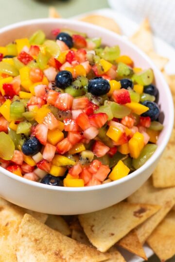 A bowl of fresh fruit salsa made with diced strawberries, mango, kiwi, and blueberries, served with a side of cinnamon sugar tortilla chips.
