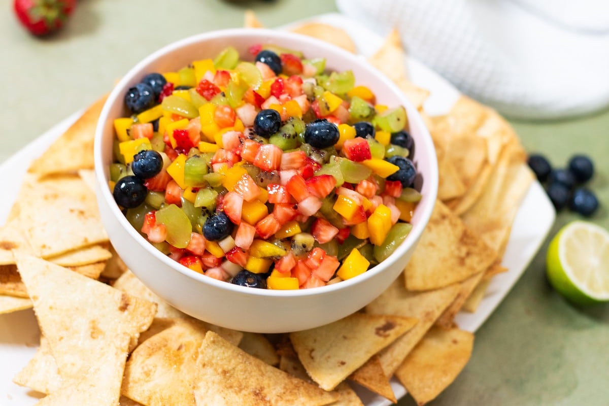 A bowl of fresh fruit salsa made with diced strawberries, mango, kiwi, and blueberries, served with a side of cinnamon sugar tortilla chips.