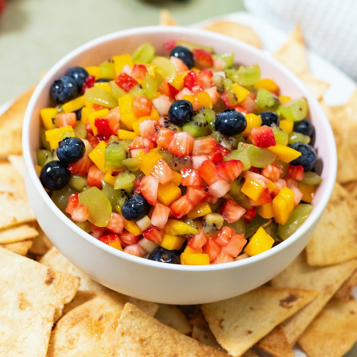 A white bowl filled with fresh fruit salsa made of diced strawberries, mango, blueberries, and kiwi, surrounded by crispy tortilla chips.