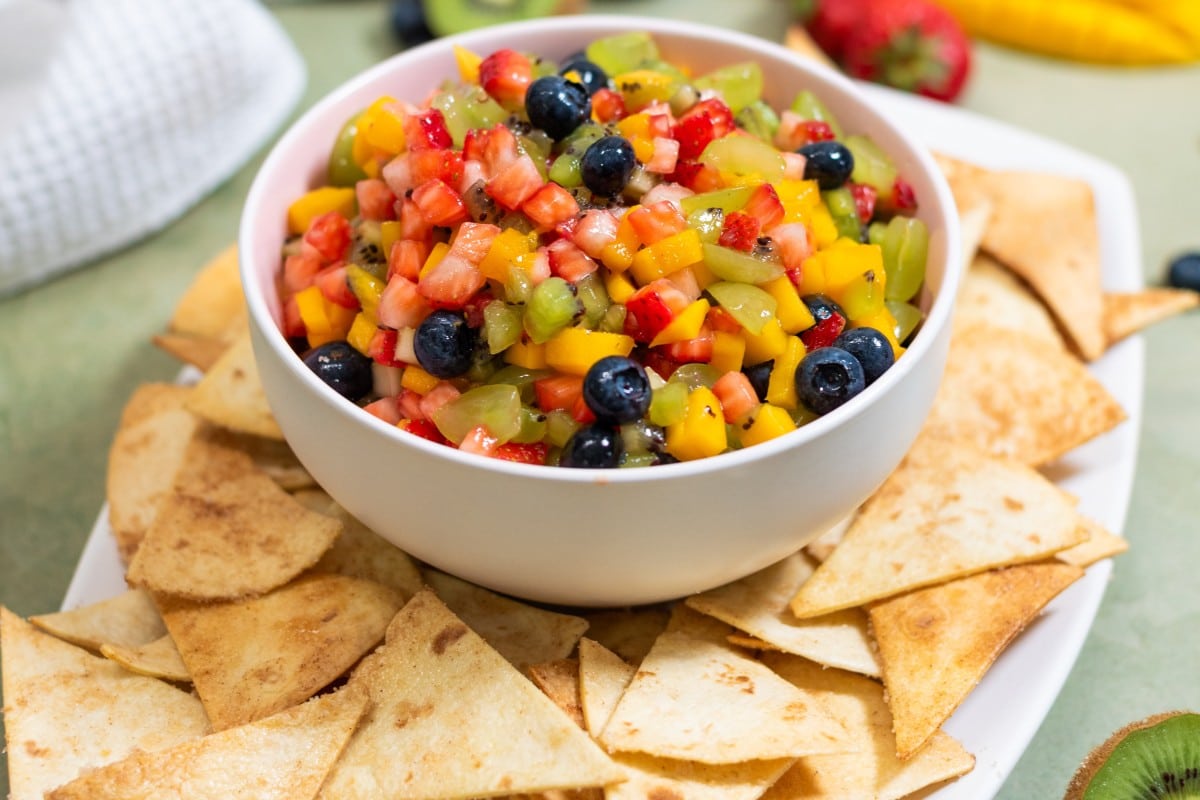 A white bowl of fresh fruit salsa made with diced strawberries, kiwi, mango, and blueberries, served with crispy cinnamon sugar tortilla chips.