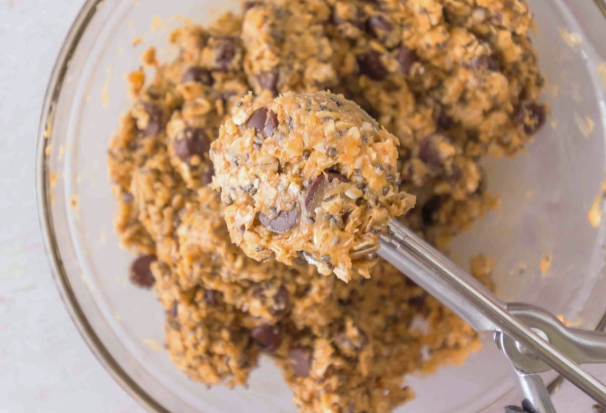 A close-up of oatmeal cookie dough mixture with chocolate chips in a clear glass bowl. A metal scoop is lifting a portion of the dough, perfect for baking protein balls.