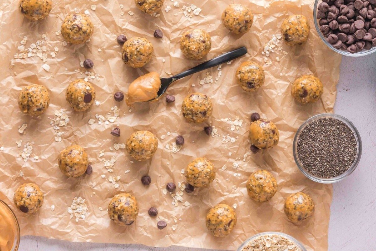 Peanut butter energy bites on parchment paper, surrounded by scattered oats, chocolate chips, chia seeds, a spoon with peanut butter, and small bowls of ingredients—perfect for oatmeal protein balls.