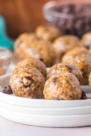 A stack of white plates holds several peanut butter oatmeal protein balls, with a bowl of peanut butter and a bowl of chocolate chips in the background.