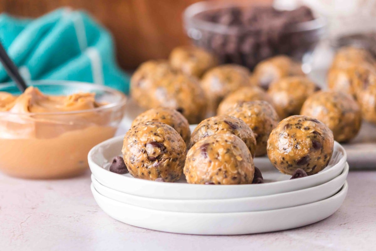 A stack of white plates holds several peanut butter oatmeal protein balls, with a bowl of peanut butter and a bowl of chocolate chips in the background.