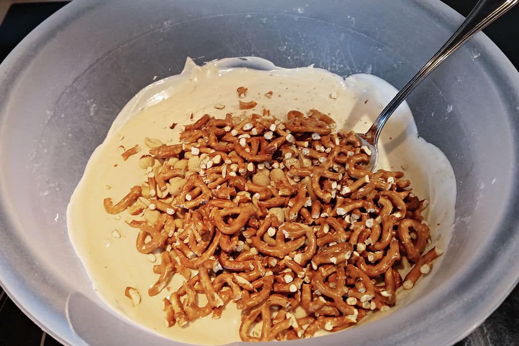 A metal spoon in a mixing bowl containing cream-colored almond bark pretzel candy batter topped with a pile of crushed pretzels.