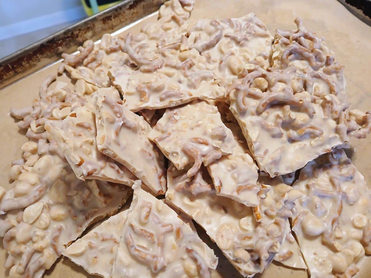 Broken pieces of white almond bark studded with pretzels and peanuts on a baking sheet.