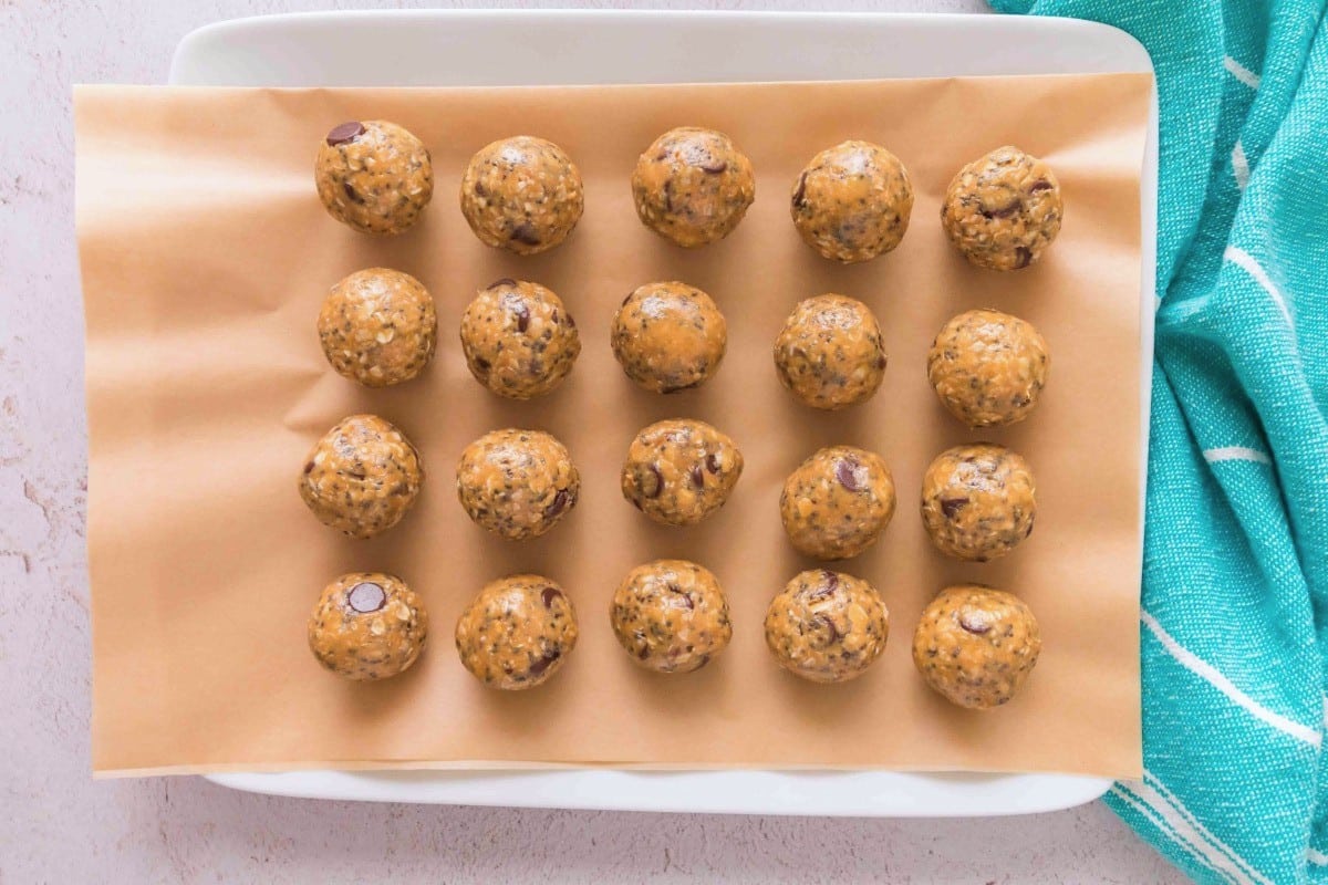 Twenty round oatmeal protein balls with chocolate chips are arranged in neat rows on parchment paper atop a rectangular white tray, next to a blue and white towel, ready to boost your energy.