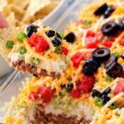 A hand holds a tortilla chip scooped into a Mexican 7 layer taco dip topped with black olives, diced tomatoes, shredded cheese, and green onions.