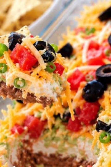 A hand holds a tortilla chip scooped into a Mexican 7 layer taco dip topped with black olives, diced tomatoes, shredded cheese, and green onions.