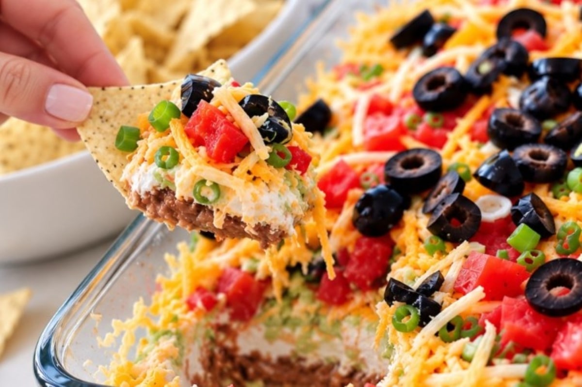 A hand holds a tortilla chip scooped into a Mexican 7 layer taco dip topped with black olives, diced tomatoes, shredded cheese, and green onions.