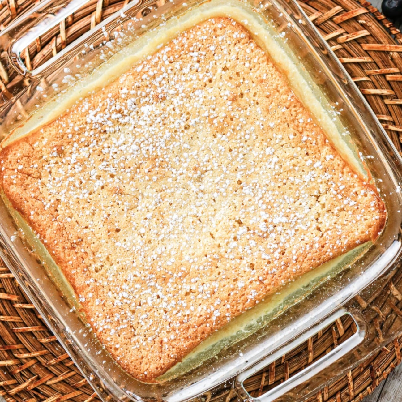 A square pudding cake topped with powdered sugar in a glass baking dish.