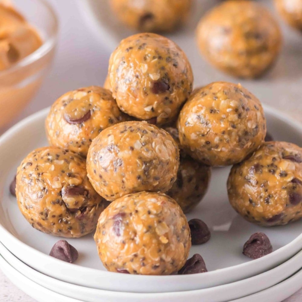 A stack of round protein balls made with oats, peanut butter, chocolate chips, and chia seeds on a white plate, with extra chocolate chips scattered nearby.