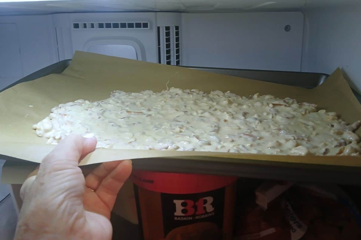 A hand holds a baking sheet of almond bark pretzel candy covered in white chocolate inside a freezer above a Baskin-Robbins container.