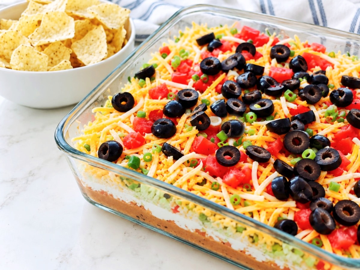 A glass baking dish containing a Mexican 7 layer taco dip topped with shredded cheese, diced tomatoes, sliced black olives, and green onions, served with a bowl of tortilla chips.