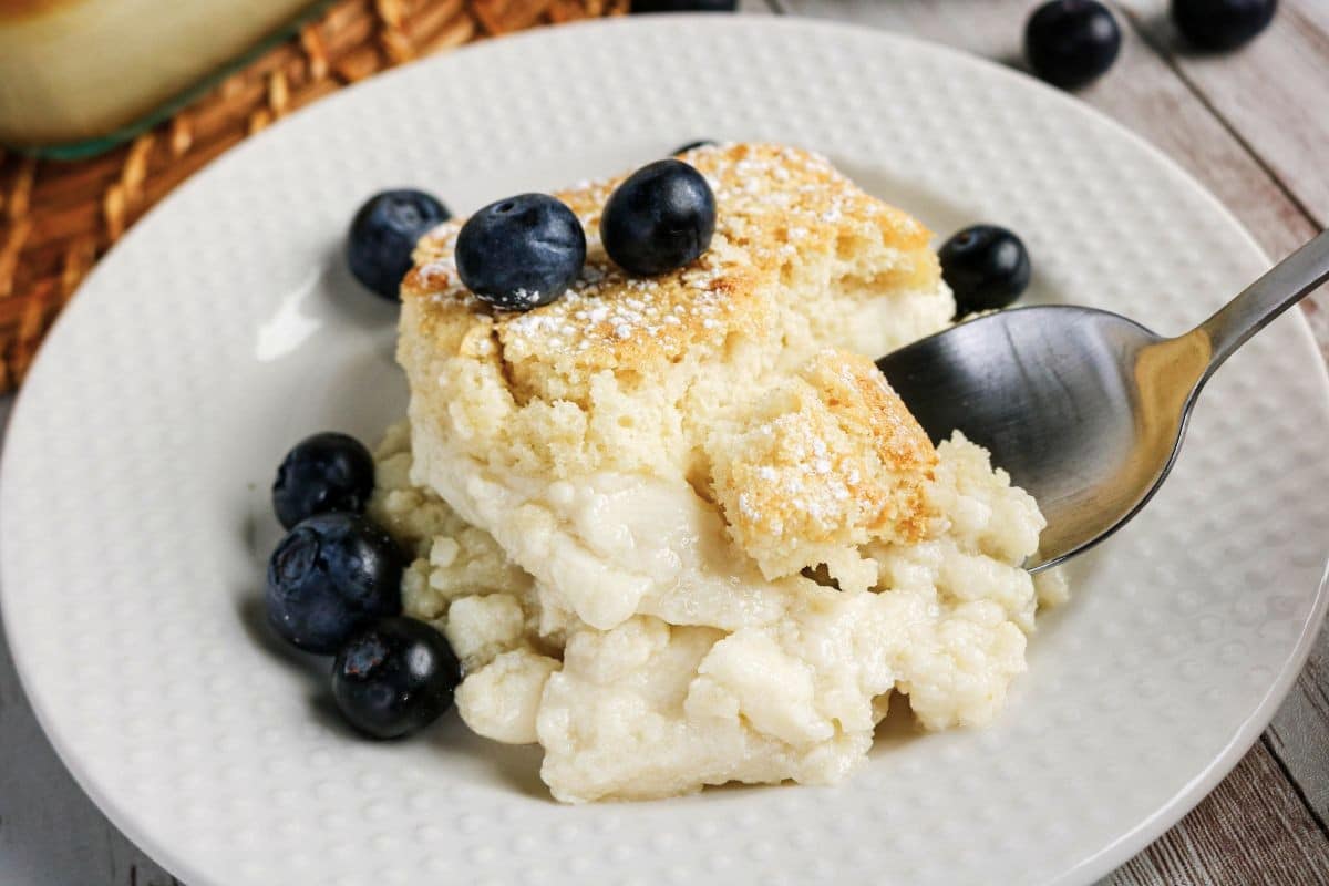 A spoonful of creamy blueberry cobbler dessert on a white plate, garnished with fresh blueberries and powdered sugar.